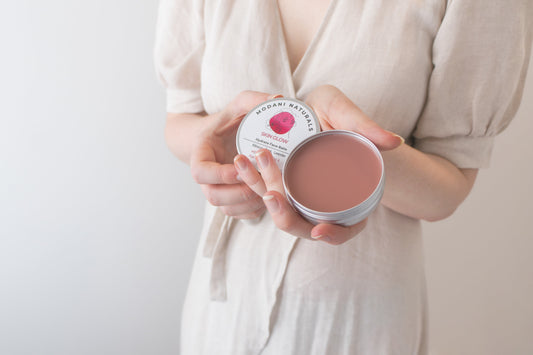 Modani Naturals Skin Glow Hydrate Balm with rosy tint, held in hands, open tin showing creamy texture.