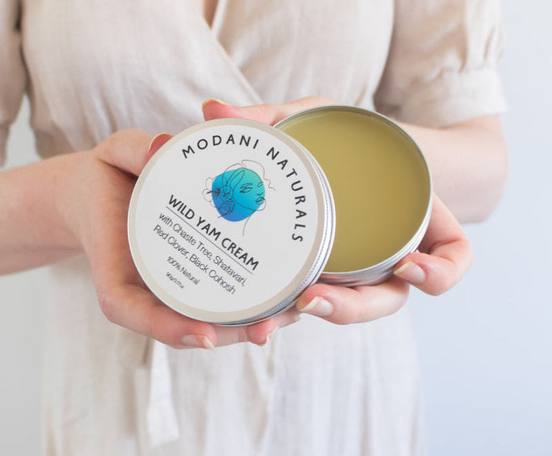 Woman wearing pale pink dress holding open tin of Modani Naturals Wild Yam Cream showing the product inside on light coloured serene background.