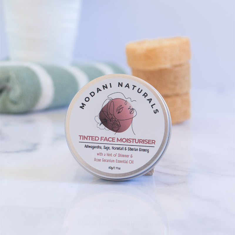 Modani Naturals Tinted Face Moisturiser, featuring a container with a label that includes a illustration of a face and ashwagandha, sage, horsetail and rose geranium essential oil on marble surface with 3 soaps in the background. .