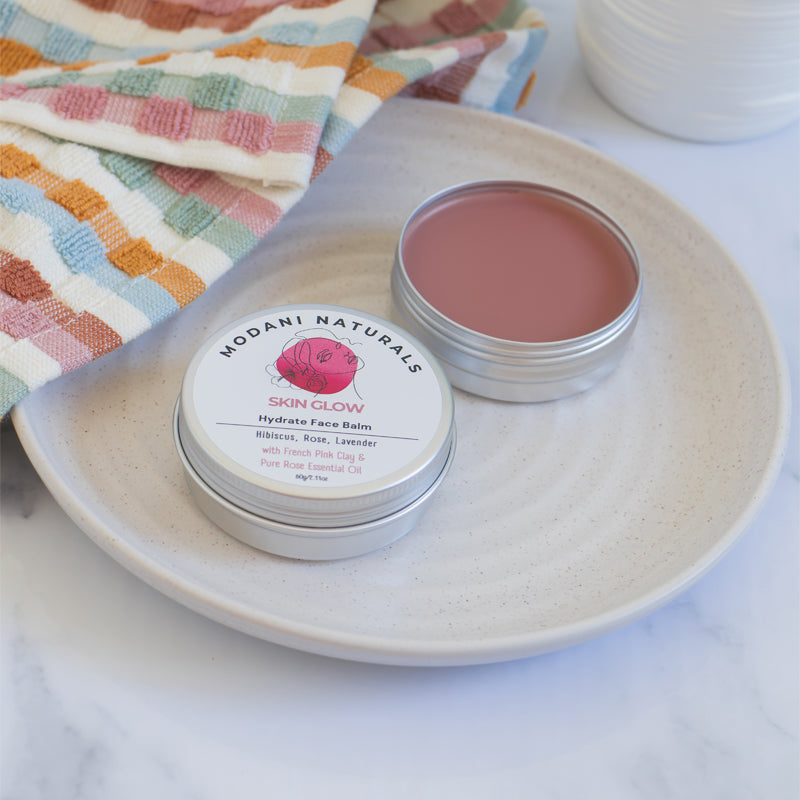Open tin of Modani Naturals Skin Glow Hydrate Face Balm with Hibiscus, Rose, Lavender showing colour and texture of contents displayed on ceramic dish with colourful towel on the side.