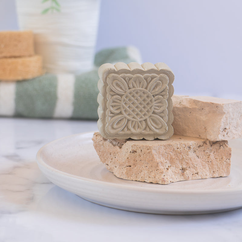 Green French Clay body cleanse and mask bar with intricate floral pattern by 'Modani Naturals' placed on stone pedestal in a ceramic dish on a white background with towel and white vase.