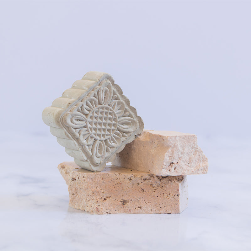 Green French Clay body cleanse and mask bar with intricate floral pattern by 'Modani Naturals' placed on stone pedestal on a white background