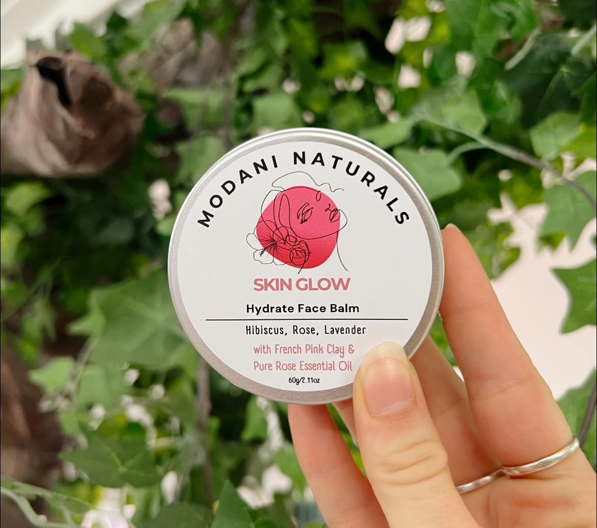 Woman's hand holding a tin of Modani Naturals Skin Glow Hydrate Face Balm with Hibiscus, Rose, Lavender with green foliage in background.