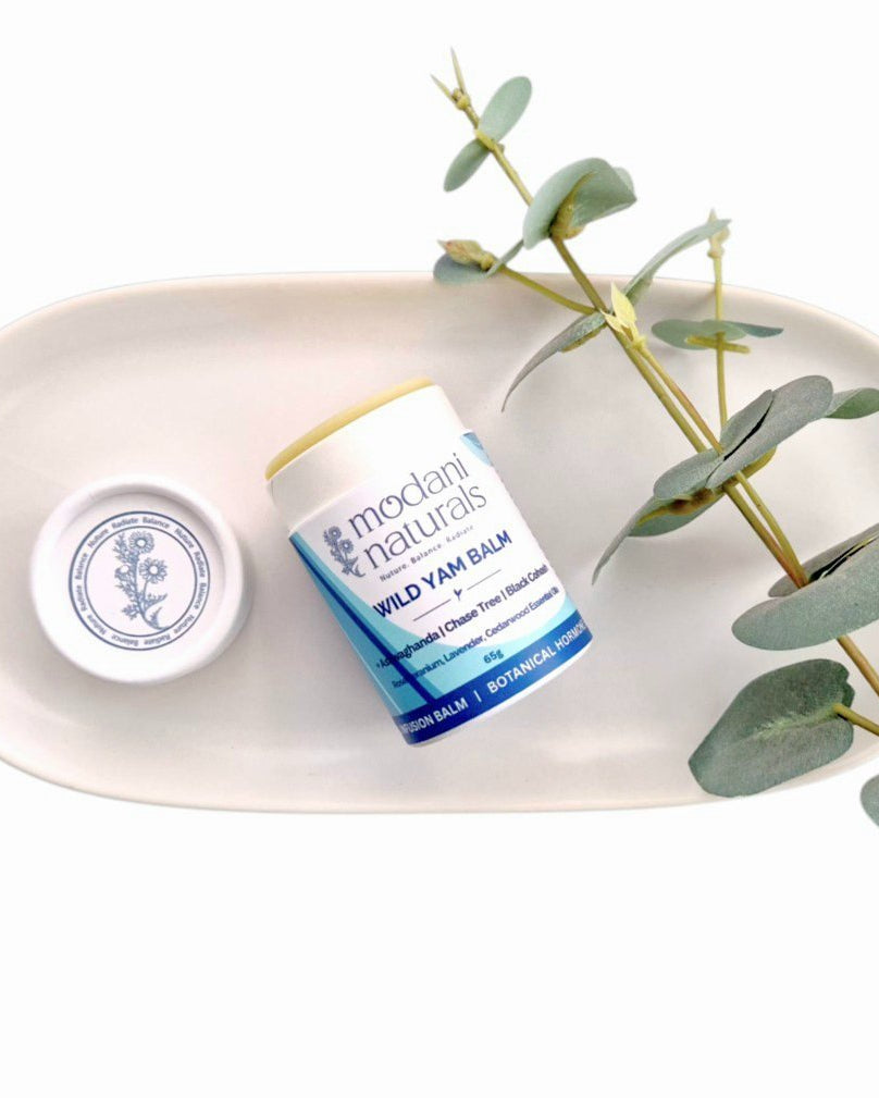 A white oval tray on a white background holding a small white jar labeled 'modani naturals WILD YAM BALM' with its lid placed beside it, styled with a green eucalyptus branch.